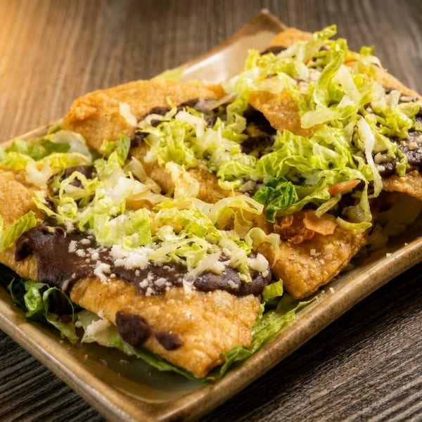 Golden fried tacos topped with shredded lettuce, dark sauce, and crumbled white cheese on a plate.