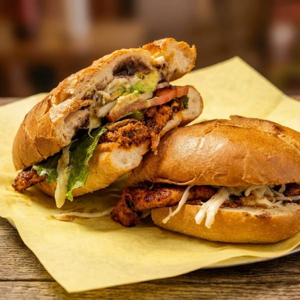 Two delicious-looking tortas, one open to show avocado, tomato, and meat, on yellow paper.